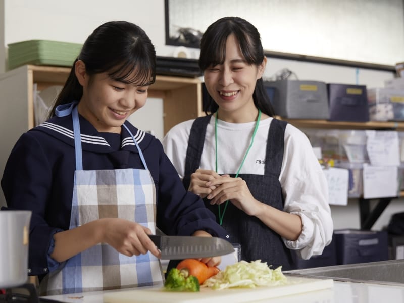 スタッフに見守られながら、笑顔で野菜を切る制服姿の生徒