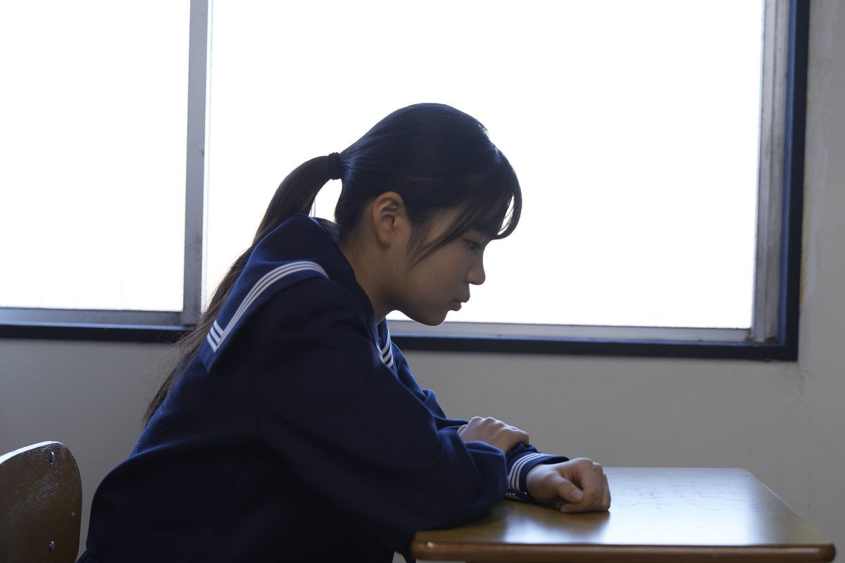 教室の窓辺の机で、うつむいて座る制服姿の生徒